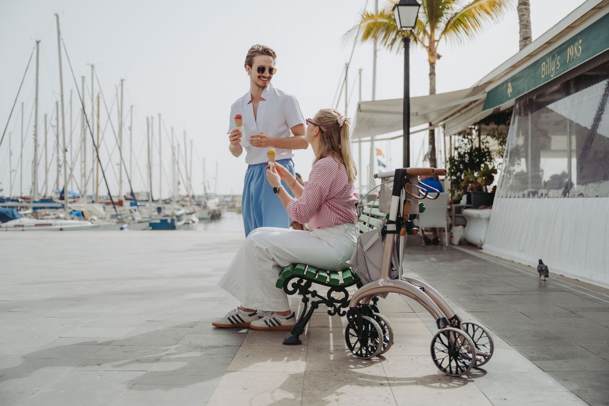 Ein Paar ist Eis auf einer Bank, daneben steht ein Rollator in einem creme Farbton.