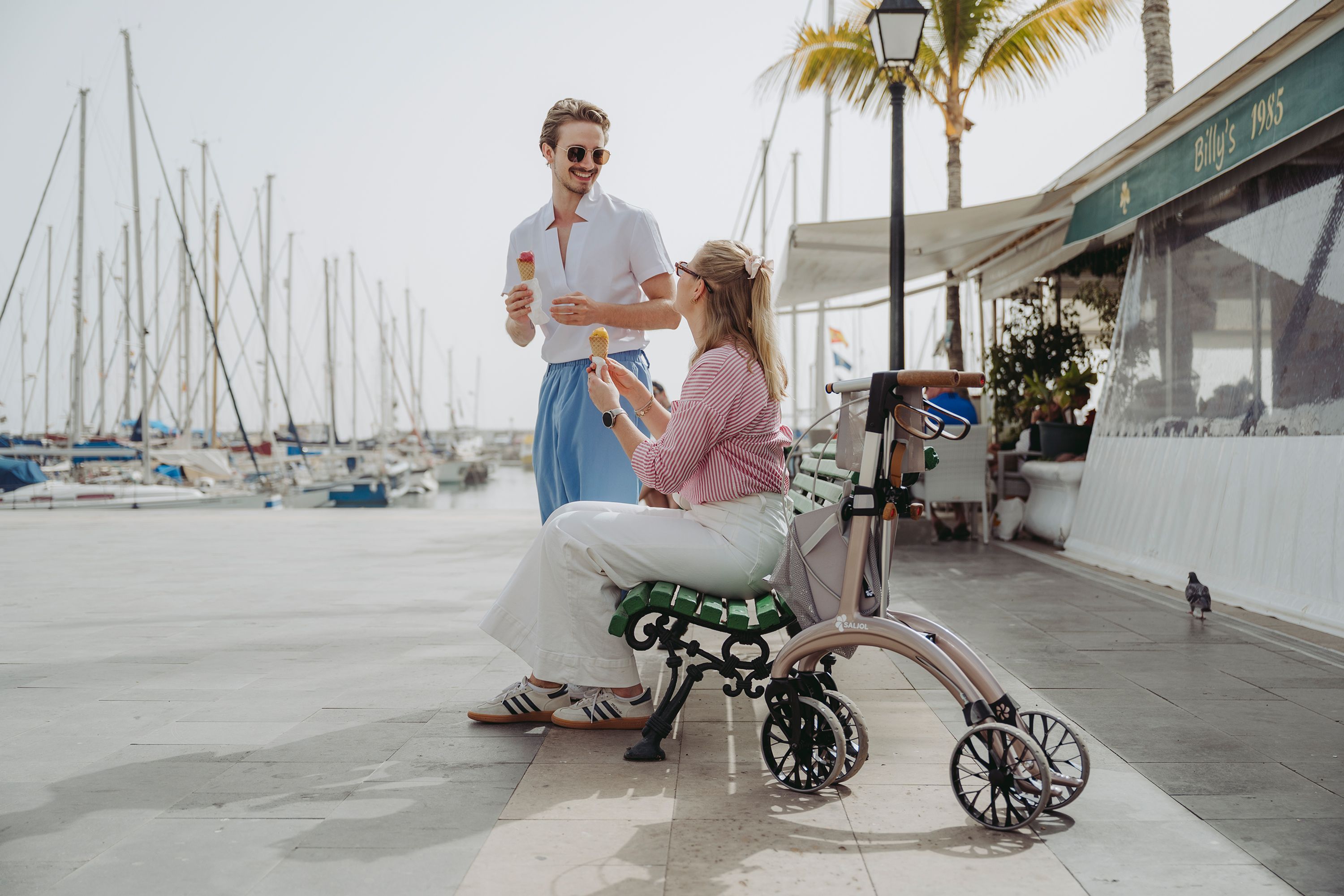 Ein Paar ist Eis auf einer Bank, daneben steht ein Rollator in einem creme Farbton.